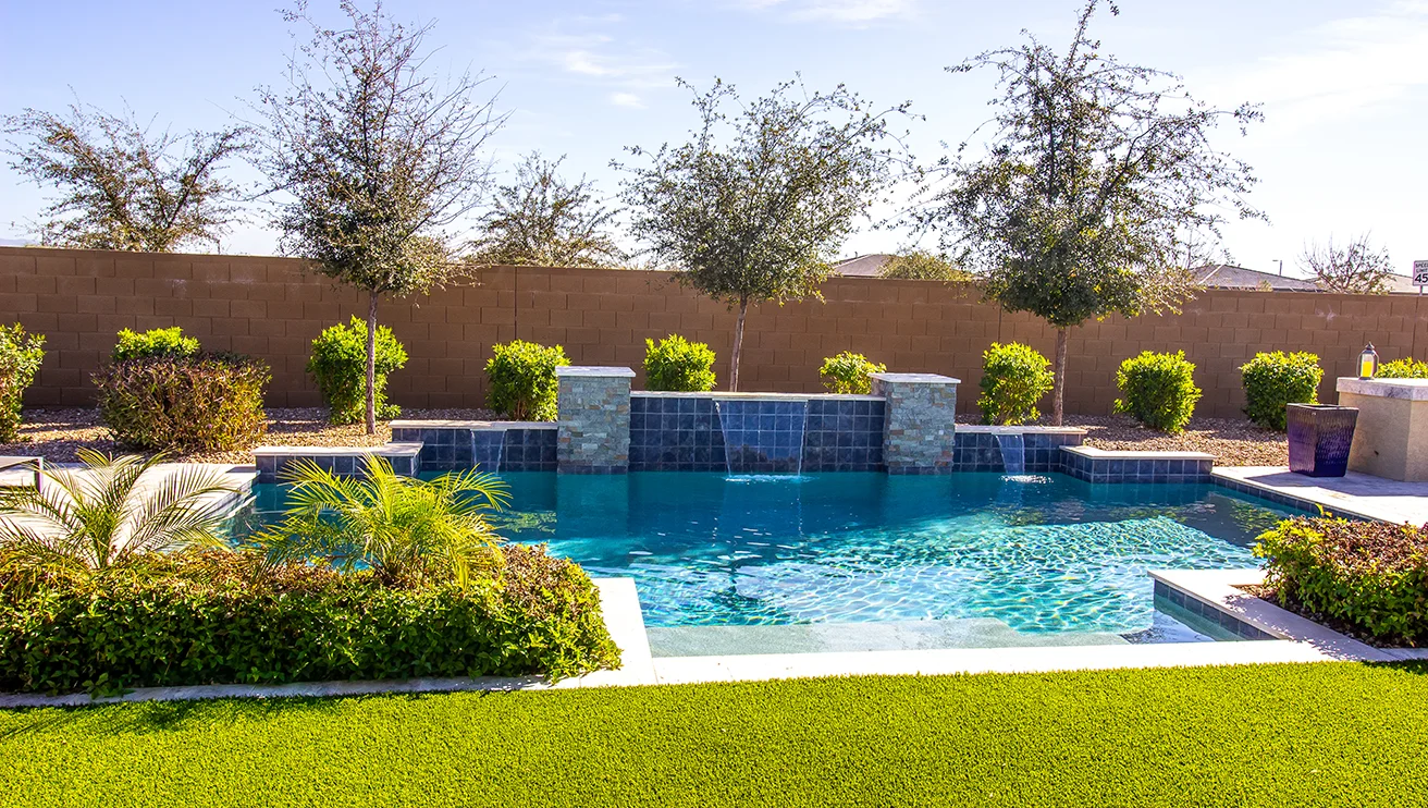 An inground swimming pool with built-in waterfalls, bordered by shrubs and trees.